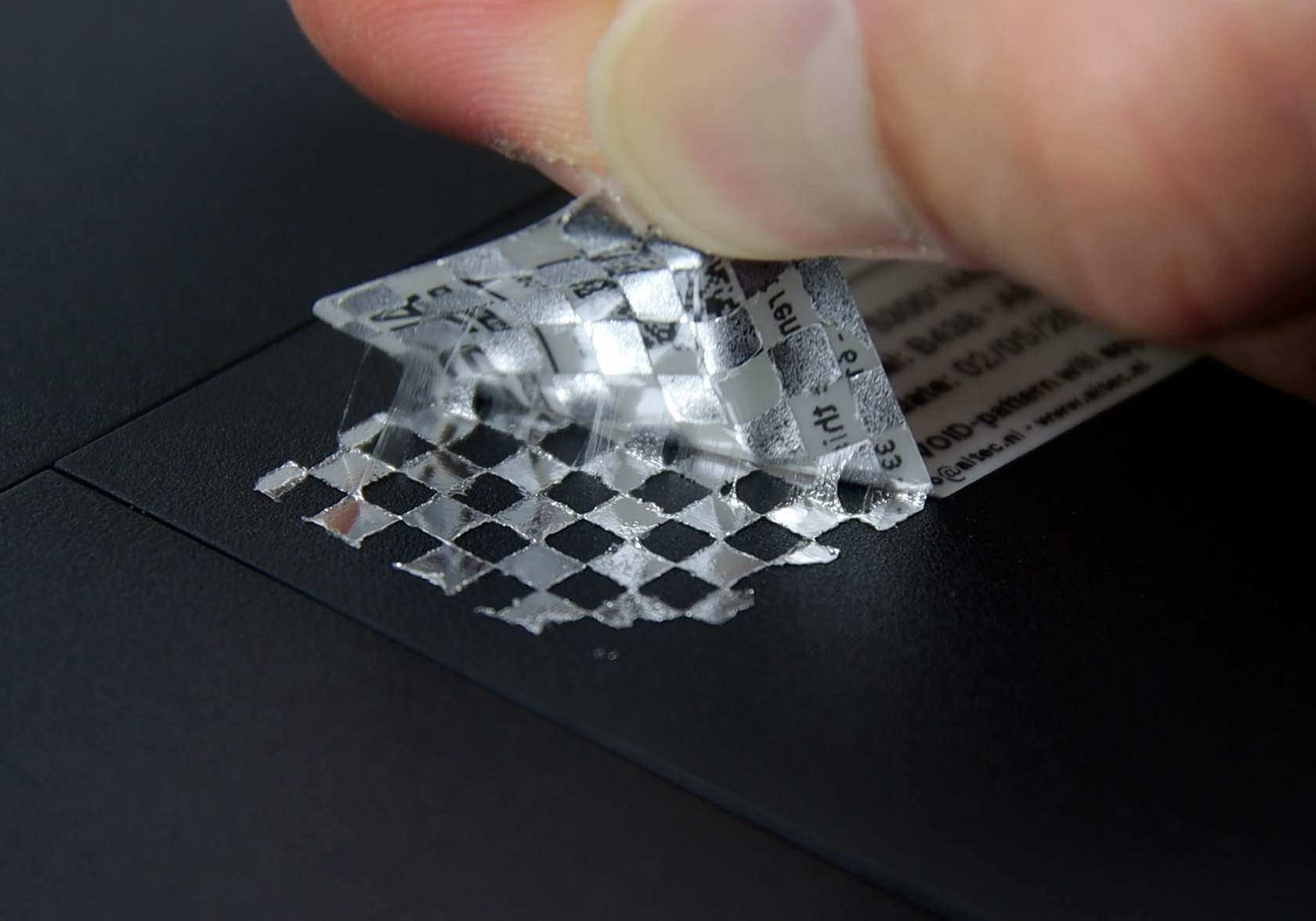 Close-up van een hand die een fraudebestendig label verwijdert, waarbij een schaakbordpatroon op de ondergrond zichtbaar wordt